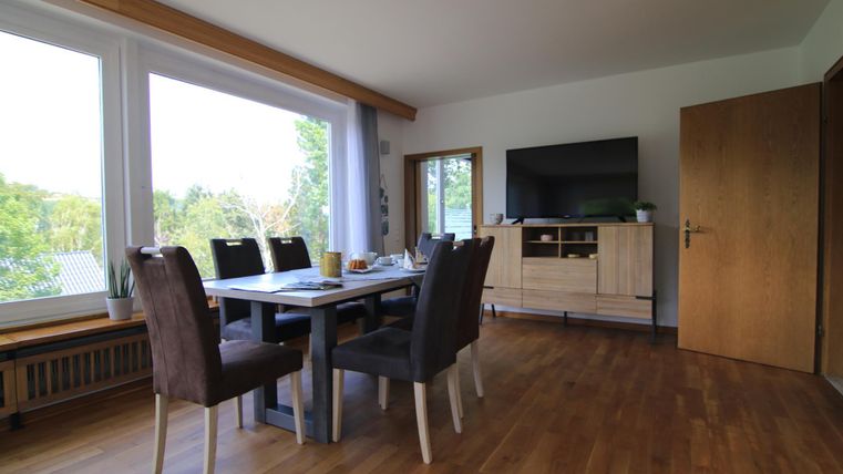 Ein helles Wohnzimmer mit einem Esstisch und Stühlen. Im Hintergrund sind große Fenster und ein Fernseher zu sehen.