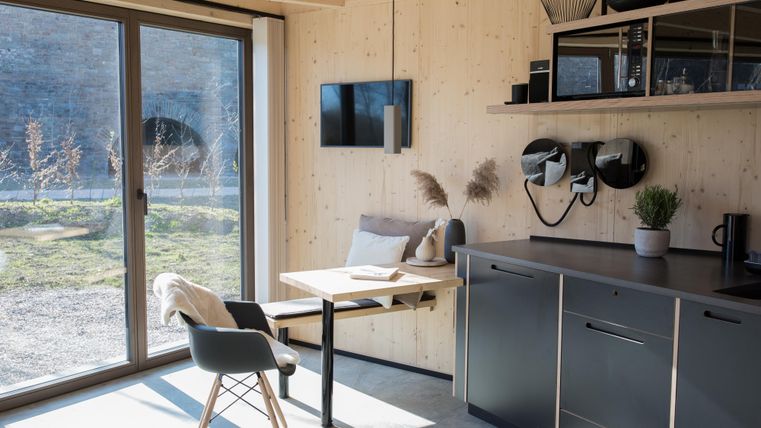 Ein modernes, helles Zimmer mit Holzverkleidung und großen Fenstern. Es gibt einen kleinen Tisch, Stühle und eine gut ausgestattete Küchenzeile.