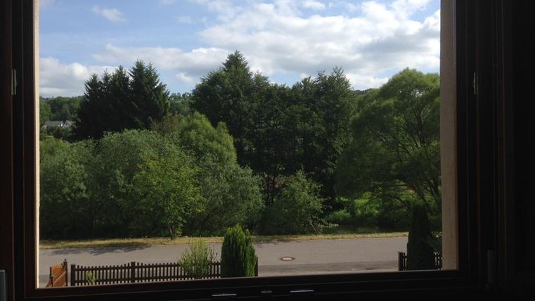 Ein Blick aus einem Fenster auf eine grüne Landschaft mit Bäumen und einem klaren Himmel. Die Szenerie vermittelt eine ruhige und natürliche Atmosphäre.