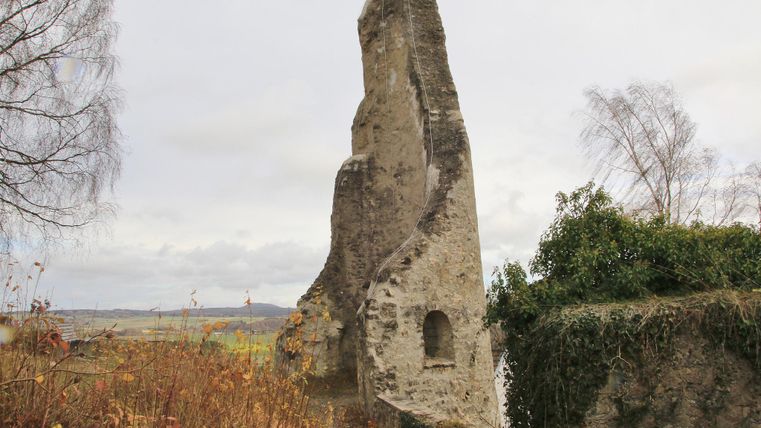 Eine alte Ruine steht inmitten von trockenem Gras und Sträuchern. Der Himmel ist bewölkt und die Landschaft wirkt ruhig.