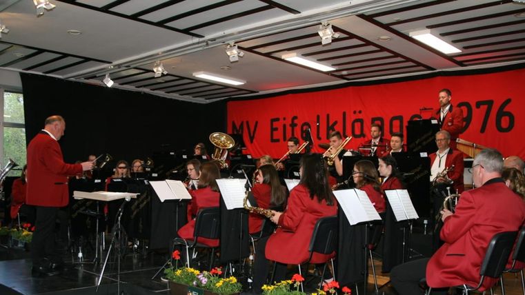 Eine Blaskapelle spielt auf einer Bühne. Die Musiker tragen rote Jacken und sind von Blumen geschmückt.