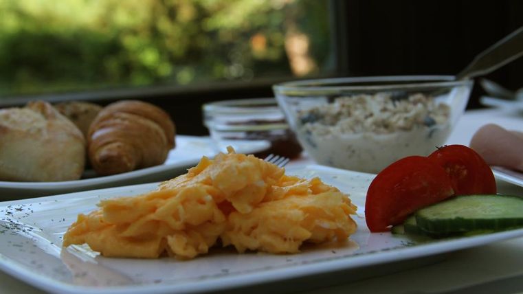 Ein Teller mit Rührei, einer Tomate und Gurkenscheiben. Im Hintergrund sind ein Croissant und eine Schüssel mit Joghurt zu sehen.