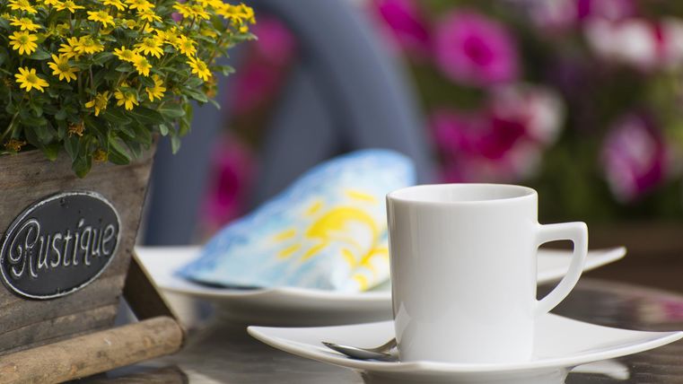 Eine weiße Tasse steht auf einem Tisch neben einem bunten Kissen und einem Blumenkübel mit gelben Blumen. Im Hintergrund sind violette Blüten zu sehen.