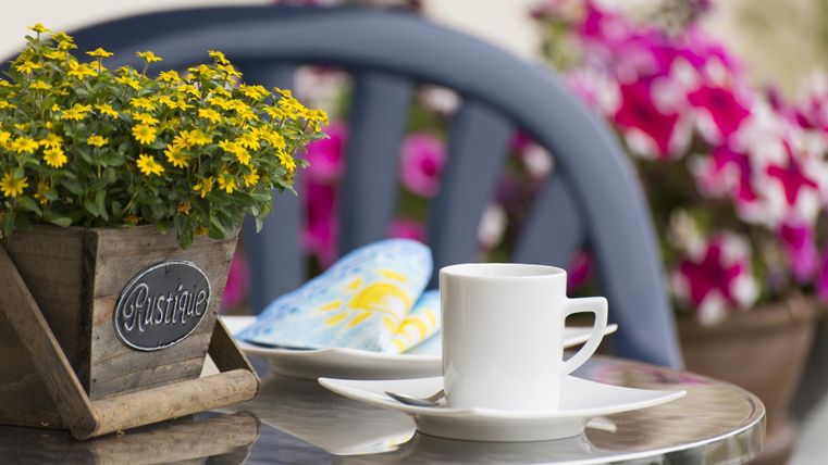 Ein Tisch im Freien mit einer Tasse Kaffee und einer kleinen Blume in einem Topf. Im Hintergrund sind bunte Blumen zu sehen.