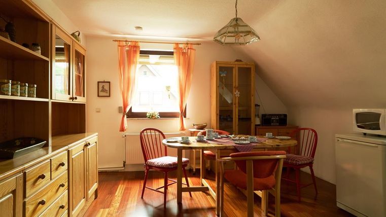 Ein gemütlicher Essbereich in einer Wohnung mit einem Holztisch und roten Stühlen. Die Fenster sind mit hellen Vorhängen geschmückt und lassen viel Tageslicht herein.