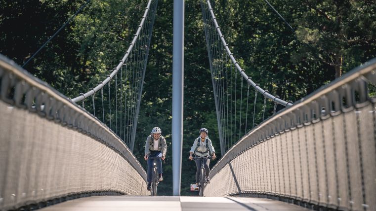 Zwei Radfahrer auf der Victor-Neels-Brücke, umgeben von Bäumen.