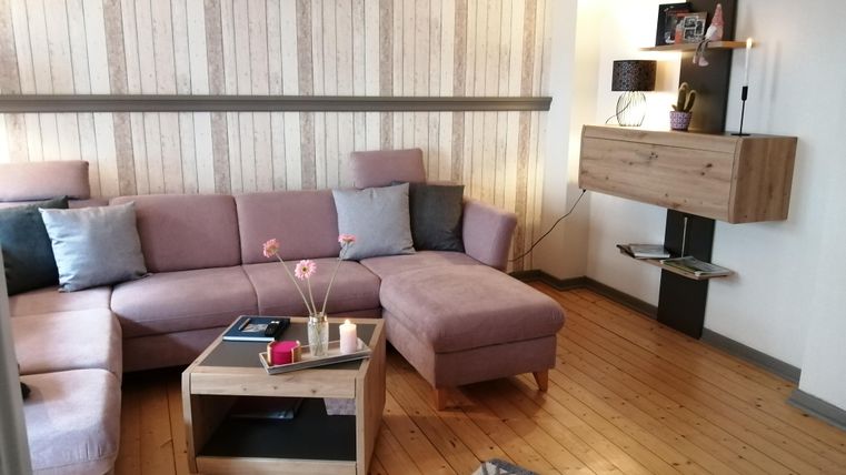 Ein gemütliches Wohnzimmer mit einem großen, lila Sofa und einem Holztisch. An der Wand befindet sich ein moderner Regal mit Pflanzen und Dekoration.