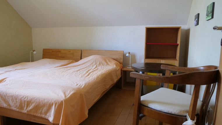 Ein einfaches Schlafzimmer mit einem Doppelbett und einer Holzkommode. Ein kleiner Tisch und ein Stuhl ergänzen die Einrichtung.