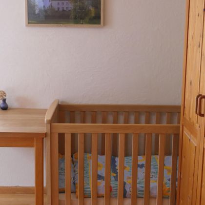 Ein helles Kinderzimmer mit einem Holzbett und einem kleinen Tisch. An der Wand hängt ein Bild eines Schlosses.