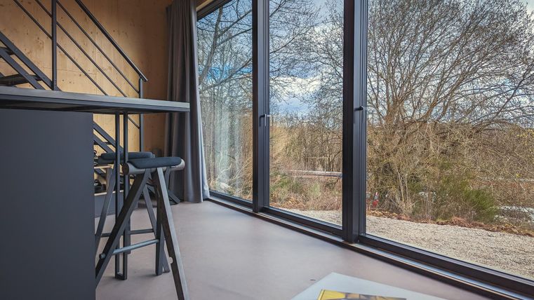 Ein modernes Zimmer mit großen Fenstern und einem Blick auf die Natur. Ein Tisch und Stühle sind im Vordergrund sichtbar, und ein Bücherstapel liegt auf dem Tisch.