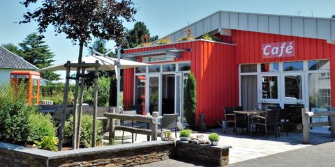 Ein einladendes Café mit buntem Äußeren in einer grünen Umgebung. Es gibt Sitzmöglichkeiten im Freien und viel Sonnenschein.