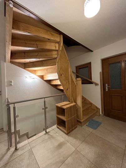 Ein einladender Eingangsbereich mit einer Holztreppe und einem großen Eingangstür. Der Boden ist mit Fliesen ausgelegt und der Raum wirkt hell und freundlich.