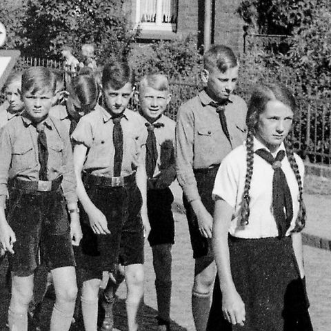 Eine Gruppe von Kindern in historischen Uniformen geht auf einer Straße. Sie tragen Hemden mit Kravatte und Shorts.