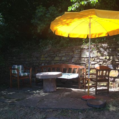 Eine gemütliche Sitzecke im Freien mit einem runden Tisch und Stühlen. Ein großer gelber Sonnenschirm spendet Schatten vor einer steinernen Mauer.