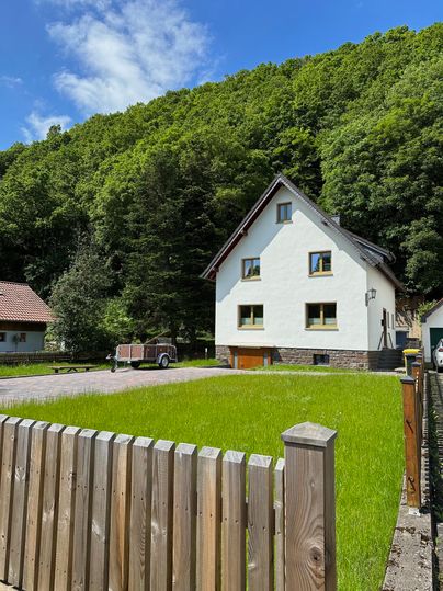 Ein weißes Haus mit grünem Rasen und einem Holzzaun. Im Hintergrund sind Bäume und ein blauer Himmel zu sehen.