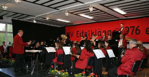 Eine Blaskapelle spielt auf einer Bühne. Die Musiker tragen rote Jacken und sind von Blumen geschmückt.