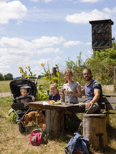 Eine Familie genießt ein Picknick an einem Tisch im Freien. Im Hintergrund sind Sonnenblumen und eine Beobachtungsstation zu sehen.