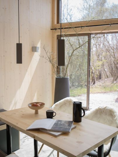 Ein modernes Esszimmer mit einem Holztisch, einer Tasse Kaffee und einem Buch. Große Fenster lassen viel natürliches Licht herein und bieten einen Blick auf die Außenlandschaft.