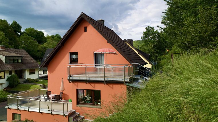 Ein hübsches, rosa Haus mit einem Balkon und einem Sonnenschirm. Umgeben von grünen Wiesen und Bäumen.