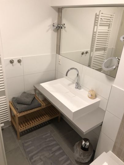 Ein modernes Badezimmer mit einer schlichten weißen Waschbeckenanlage und einem Spiegel. Handtücher sind auf einem Holzwagen ordentlich gestapelt.