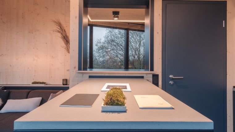 Ein moderner Raum mit einem großen Tisch und einer Fensterfront. Auf dem Tisch liegen einige Unterlagen und eine kleine Pflanze.