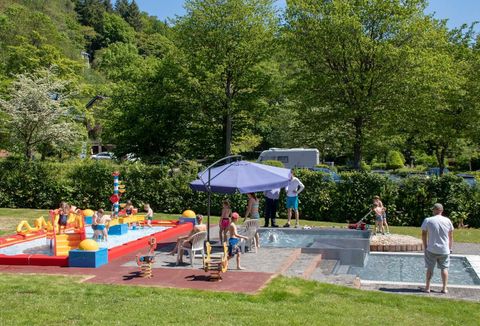 Een mooie tuin met een kinderzwembad en kleurrijk speelgoed. Op de achtergrond zijn groene bomen en enkele volwassenen te zien die ontspannen.