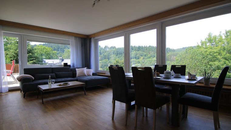 Ein heller Wohnraum mit großen Fenstern, Blick auf die grüne Landschaft und stilvoller Einrichtung. Der Raum verfügt über eine gemütliche Couch und einen Esstisch.