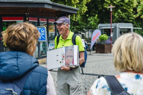 Ein älterer Mann mit einer Sonnenbrille und einem grünen Hemd hält einen Ordner in der Hand. Er spricht mit zwei Frauen in der Stadt.