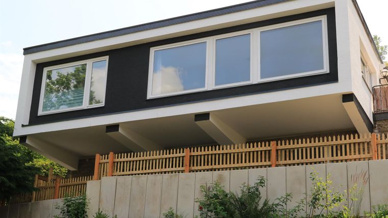 Ein modernes Haus mit einer klaren, minimalistischen Fassade. Es steht auf Pfeilern und ist von einem Holzzaun umgeben.