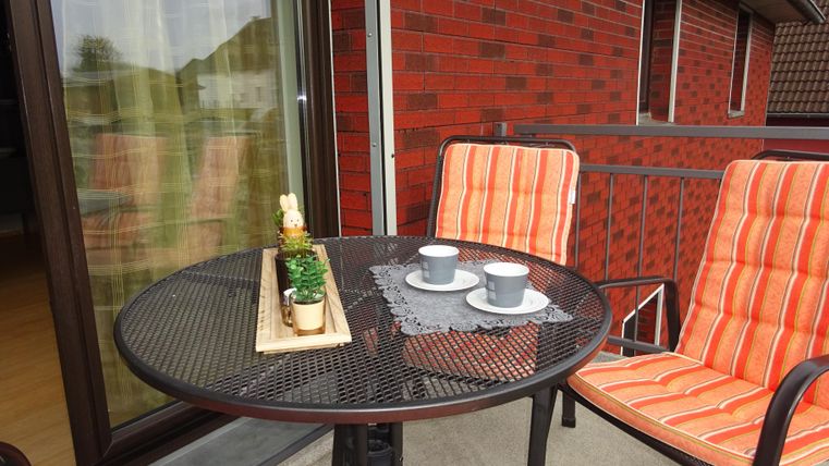 Ein kleiner Balkon mit einem runden Tisch und zwei Stühlen. Auf dem Tisch stehen zwei Tassen und eine Blumendekoration.