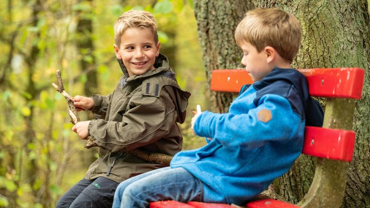 Zwei Jungen sitzen auf einer roten Bank im Wald. Der Junge links lächelt und hält einen Stock in der Hand.