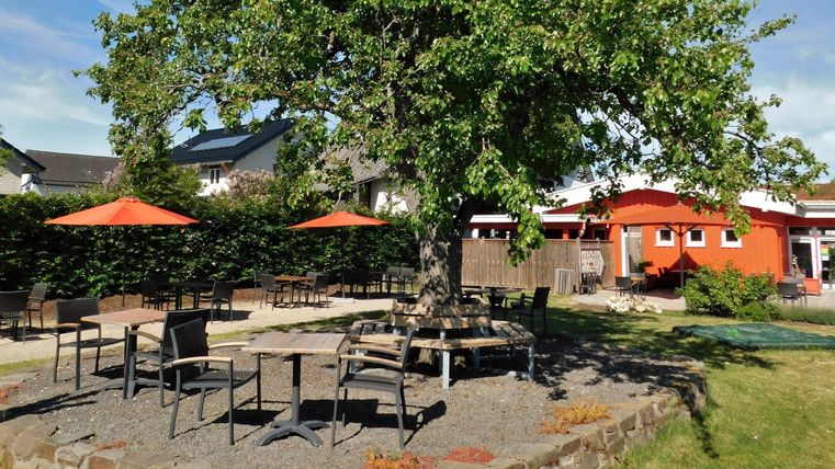 Ein einladender Außenbereich mit einem großen Baum und mehreren Tischen. Orangefarbene Sonnenschirme spenden Schatten für die Gäste.