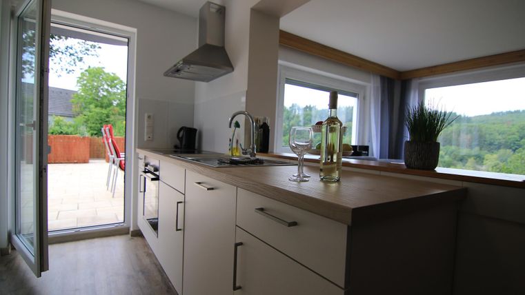 Eine moderne Küche mit einem großzügigen Fenster, das auf eine Terrasse mit Blick ins Grüne führt. Auf der Arbeitsfläche stehen Glasgefäße und Pflanzen.