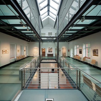 Ein moderner Innenraum mit Glasdach und mehreren Ebenen. An den Wänden hängen verschiedene Kunstwerke, während im Vordergrund Sitzgelegenheiten platziert sind.