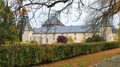 Ein historisches Gebäude, umgeben von buntem Laub und einer grünen Hecke. Der Himmel ist bewölkt, was eine herbstliche Stimmung erzeugt.