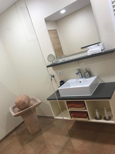 Ein modernes Badezimmer mit einem quadratischen Waschbecken und einer großen Spiegelfläche. Ein stilvoller Behälter aus Ton steht neben dem Waschbecken auf einem Regal.