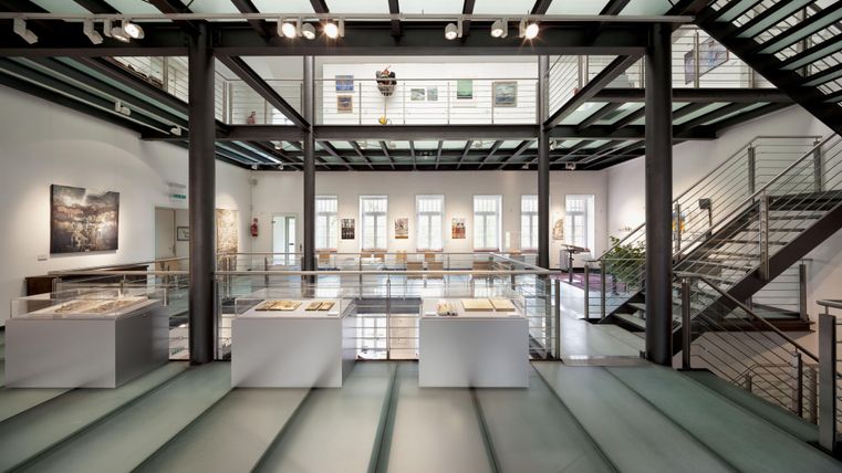 Ein modernes Ausstellungsraum mit großen Fenstern und einladender Architektur. Im Vordergrund stehen mehrere Informationsstände auf einem glatten Boden.