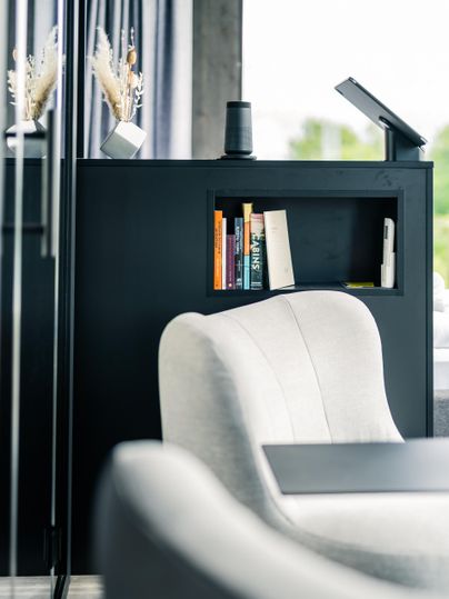 Eine moderne Wohnzimmereinrichtung mit einem eleganten, weißen Sessel und einem schwarzen Regal. Im Regal befinden sich Bücher und dekorative Elemente, während eine Lampe auf einem Tisch steht.