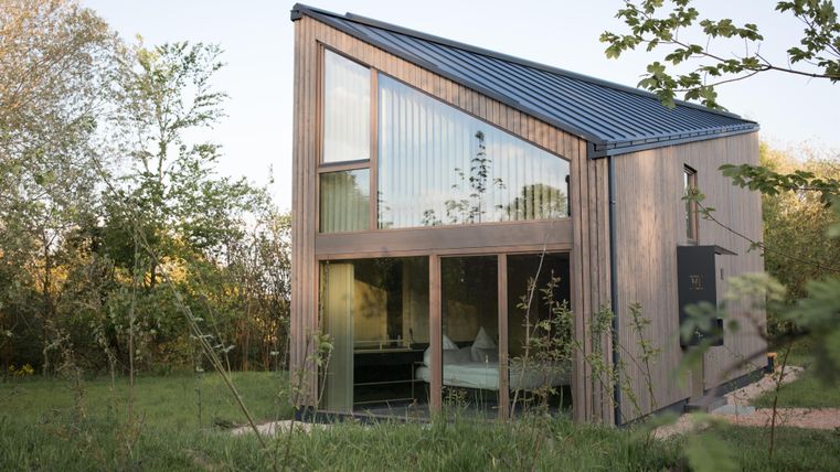 Ein modernes Holzhaus mit großen Fenstern steht in einer grünen Umgebung. Die Architektur ist stilvoll und harmonisch in die Natur integriert.