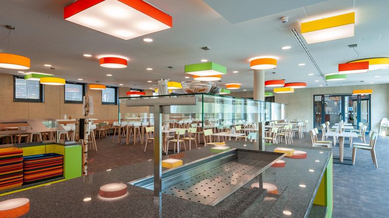 Ein modernes Restaurant mit bunten Deckenleuchten und einem hellen, einladenden Ambiente. Es gibt viele Tische und Sitzgelegenheiten für Gäste.