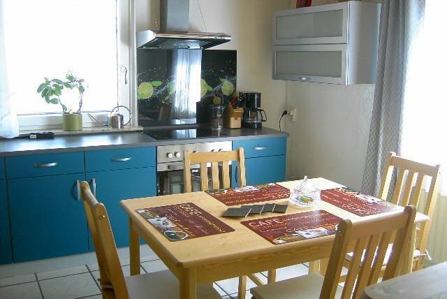 Eine moderne Küche mit blauen Schränken und einem Esstisch. Auf dem Tisch liegen dekorative Platzsets und es gibt eine Pflanze auf der Fensterbank.