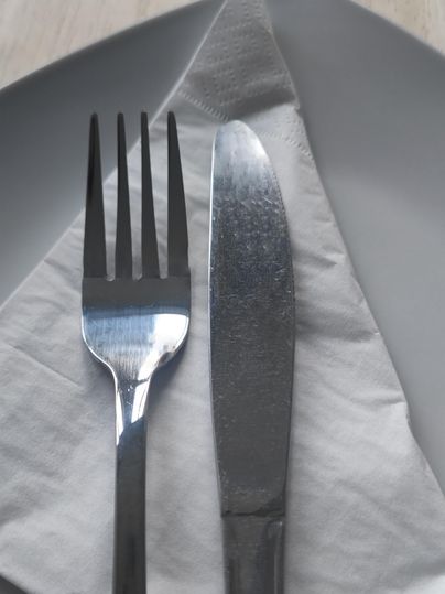 Eine Gabel und ein Messer liegen auf einer Stoffserviette auf einem grauen Teller. Die Utensilien sind aus glänzendem Edelstahl.