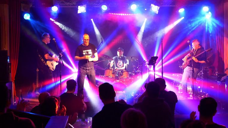 Eine Band spielt live auf einer Bühne mit bunten Lichtern. Das Publikum schaut begeistert zu und applaudiert.