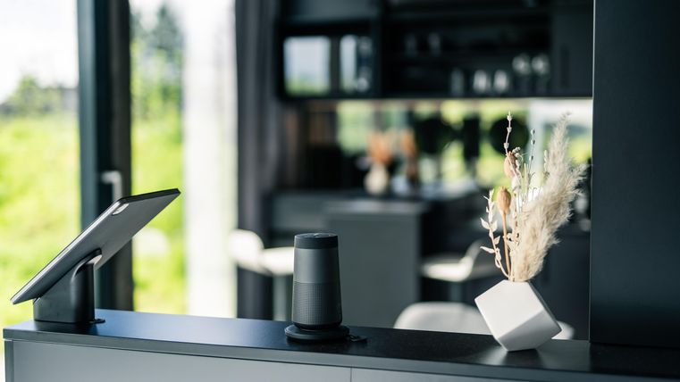 Ein moderner Raum mit einem Tablet und einem Lautsprecher auf der Theke. Im Hintergrund sind einige Möbel und natürliche Lichtverhältnisse zu sehen.