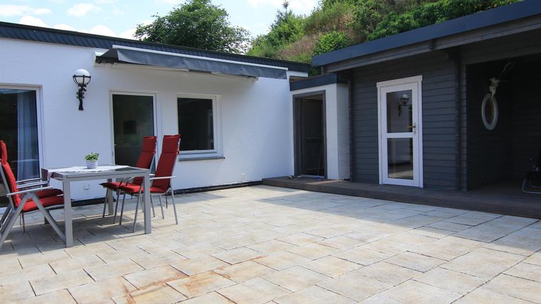 Ein schöner patio mit einem Tisch und roten Stühlen. Im Hintergrund sind zwei moderne Gebäude und etwas grüne Vegetation zu sehen.