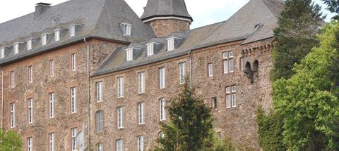 Un bâtiment historique avec un toit gris et une façade en pierre. Entouré d'arbres verts et d'un paysage pittoresque.