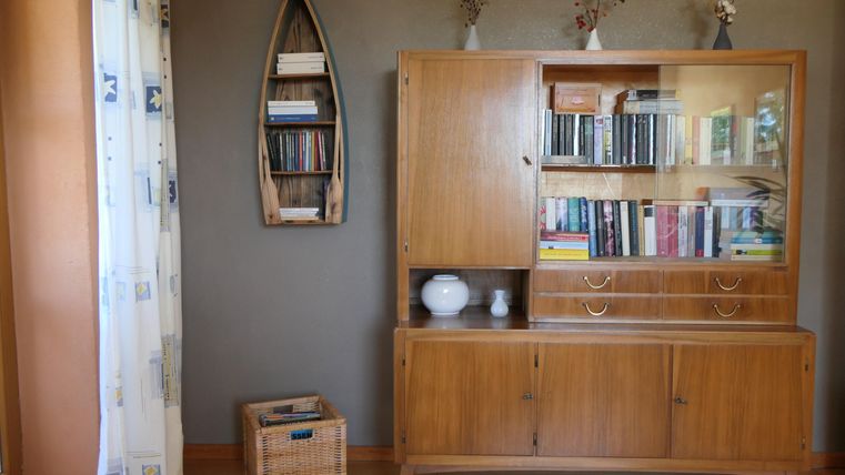 Ein gemütlicher Raum mit einem Holzschrank, der Bücher enthält. An der Wand hängt ein Regal in Form eines Bootes, und es gibt dekorative Elemente sowie einen Wäschekorb.
