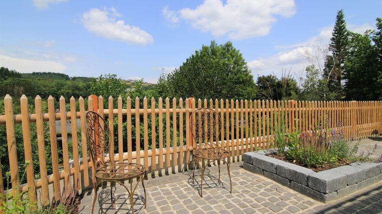 Eine gemütliche Sitzgelegenheit mit zwei Stühlen aus Metall vor einem Holzlattenzaun. Im Hintergrund sind Bäume und ein blauer Himmel zu sehen.