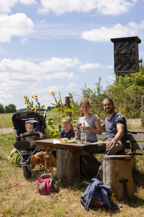 Eine Familie genießt ein Picknick an einem Tisch im Freien. Im Hintergrund sind Sonnenblumen und eine Beobachtungsstation zu sehen.