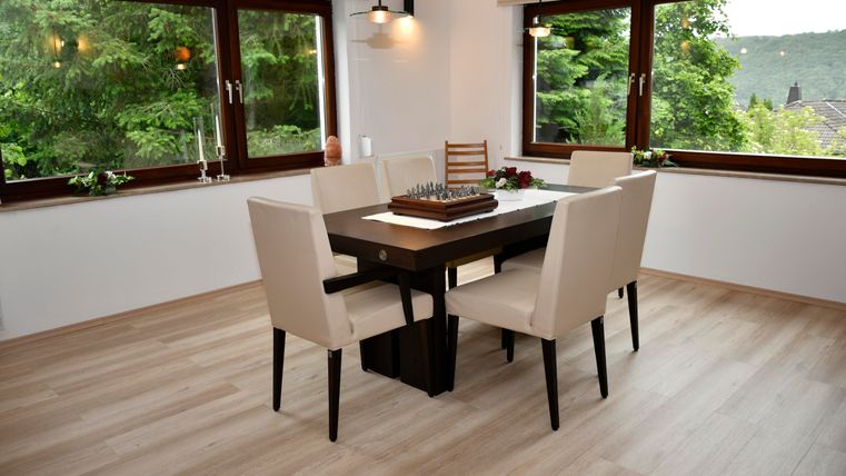 Ein heller Essbereich mit einem Tisch und sechs Stühlen. Große Fenster bieten einen Blick auf die grüne Umgebung.